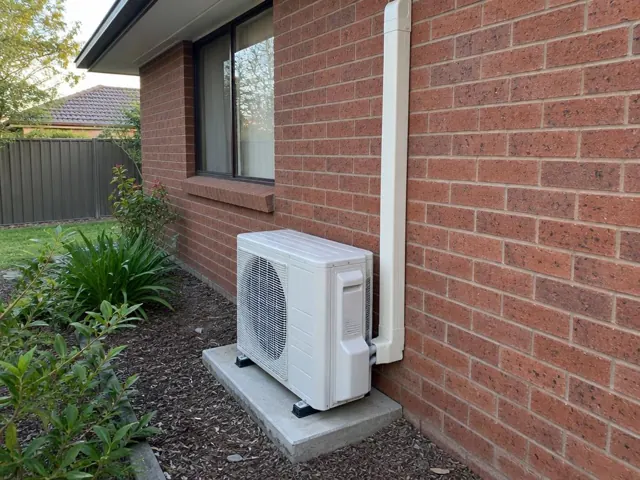 Outdoor air conditioner unit on a concrete pad beside a brick house wall in Happy Valley